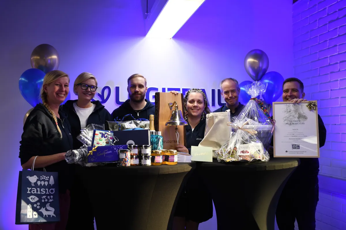 Kuusi Logitrailin johtoryhmän jäsentä hymyilee pöydän takana, joka on täynnä lahjoja, kuten laivakello ja kehystetty ikimetsä-lahjakirja.